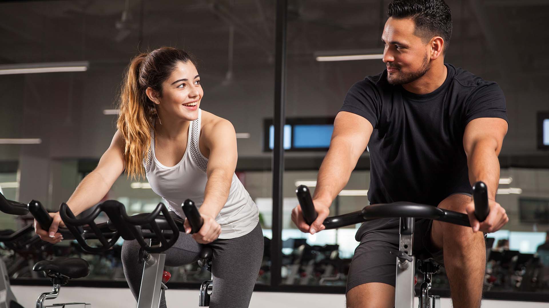 Brunette man and woman conversing while smiling at each other at the gym. They are both on the spin bikes.